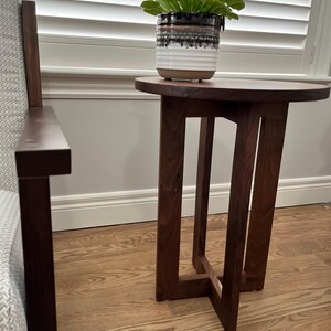 Op de afbeelding: Een donkerbruine houten bijzettafel met een rond blad en een eenvoudig, modern ontwerp. De tafel heeft &eacute;&eacute;n plank en wordt ondersteund door vier poten.