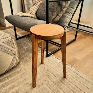 May include: A small, round wooden stool with three legs. The stool is made of light brown wood and has a simple, minimalist design. The stool is placed in front of a modern grey chair with a black metal frame. The chair has pillows and a blanket.