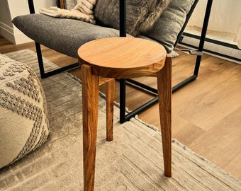 May include: A small, round wooden stool with three legs. The stool is made of light brown wood and has a simple, minimalist design. The stool is placed in front of a modern gray chair with a black metal frame. The chair has pillows and a blanket.