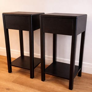 Puede incluir: Dos mesitas de noche negras con un caj&oacute;n y una balda inferior. Las mesitas tienen un dise&ntilde;o minimalista con una parte superior rectangular y una base robusta de cuatro patas. Las mesas son de madera y tienen un acabado oscuro.