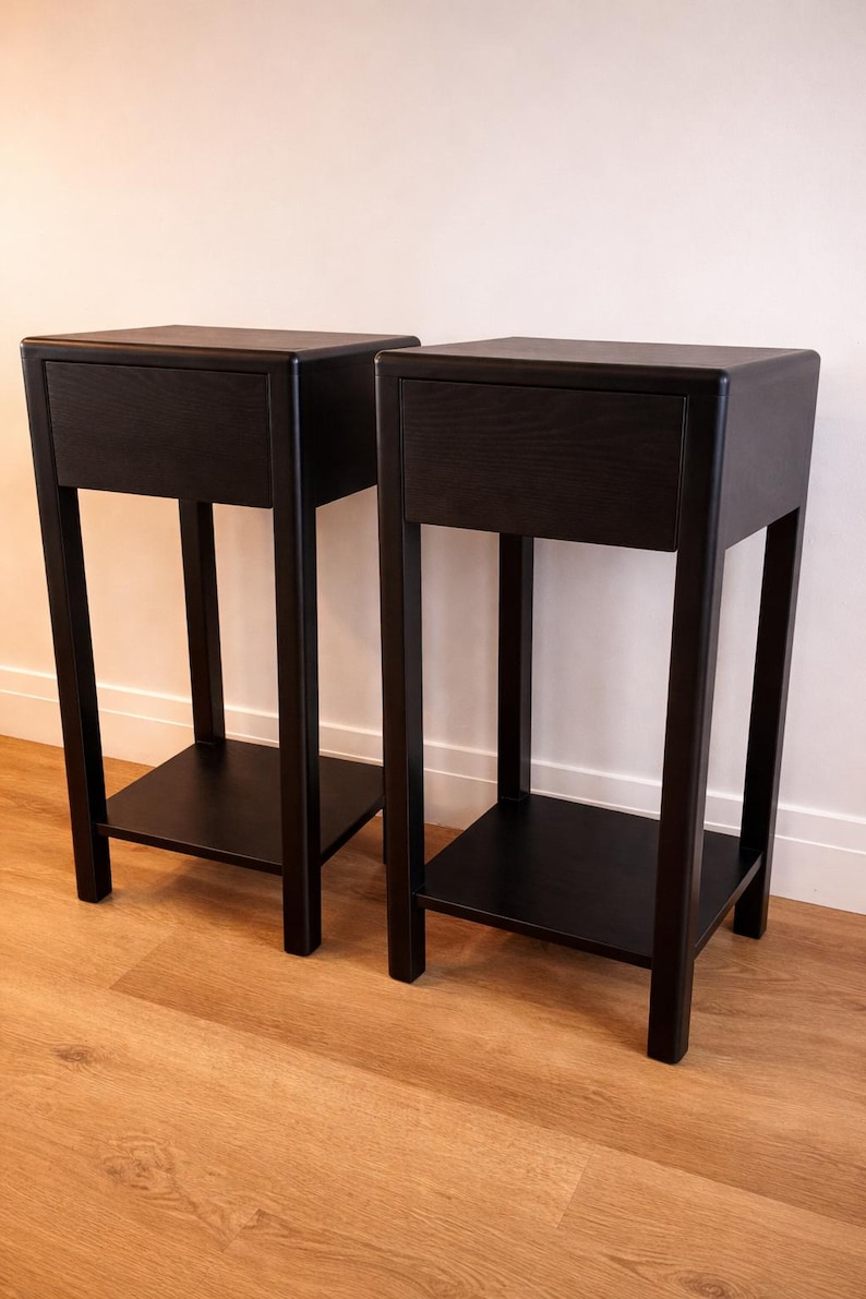 Puede incluir: Dos mesitas de noche negras con un caj&oacute;n y una balda inferior. Las mesitas tienen un dise&ntilde;o minimalista con una parte superior rectangular y una base robusta de cuatro patas. Las mesas son de madera y tienen un acabado oscuro.