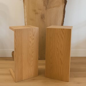 May include: Two wooden side tables with a natural wood grain finish. One table has a cantilevered top, the other is a simple rectangular block. A large wooden slab is in the background, adding to the natural aesthetic.