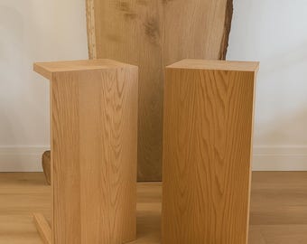 Può includere: Due tavolini in legno con finitura a venature naturali. Un tavolo ha un piano a sbalzo, l'altro è un semplice blocco rettangolare. Una grande lastra di legno è sullo sfondo, aggiungendo all'estetica naturale.