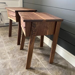 Pu&ograve; includere: Due comodini in legno con una finitura marrone scuro. Ogni comodino ha un singolo cassetto e gambe angolate. I comodini sono posizionati su un pavimento piastrellato, con un mobile bianco e una parete grigio scuro sullo sfondo.