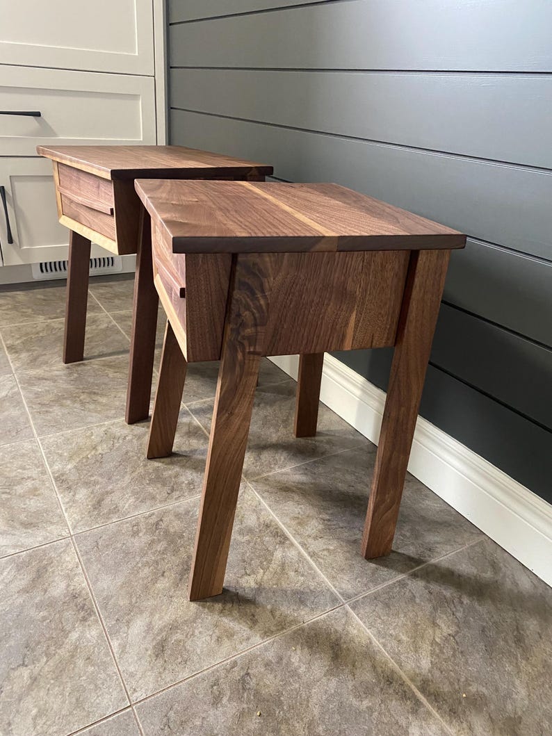Pu&ograve; includere: Due comodini in legno con una finitura marrone scuro. Ogni comodino ha un singolo cassetto e gambe angolate. I comodini sono posizionati su un pavimento piastrellato, con un mobile bianco e una parete grigio scuro sullo sfondo.