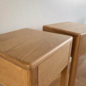 Puede incluir: Dos mesitas de noche de roble claro con esquinas redondeadas y un solo caj&oacute;n. Las mesitas tienen un acabado natural de vetas de madera y est&aacute;n sobre una pared neutra.