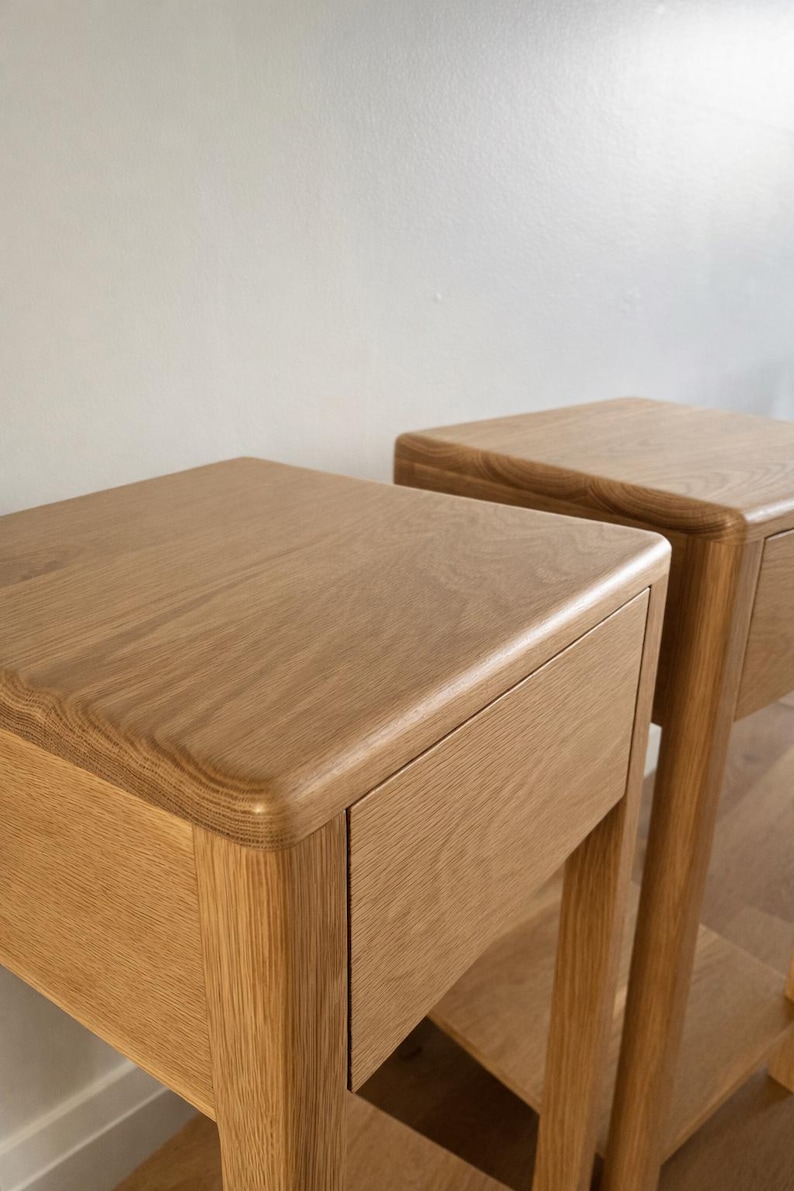 Puede incluir: Dos mesitas de noche de roble claro con esquinas redondeadas y un solo caj&oacute;n. Las mesitas tienen un acabado natural de vetas de madera y est&aacute;n sobre una pared neutra.