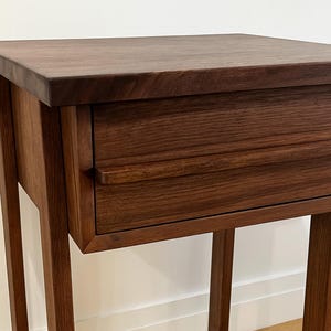 Pu&ograve; includere: Un tavolino in legno marrone scuro con un singolo cassetto. Il tavolo ha un piano rettangolare e quattro gambe sottili. Il cassetto presenta una maniglia semplice e curva. La venatura del legno &egrave; visibile.