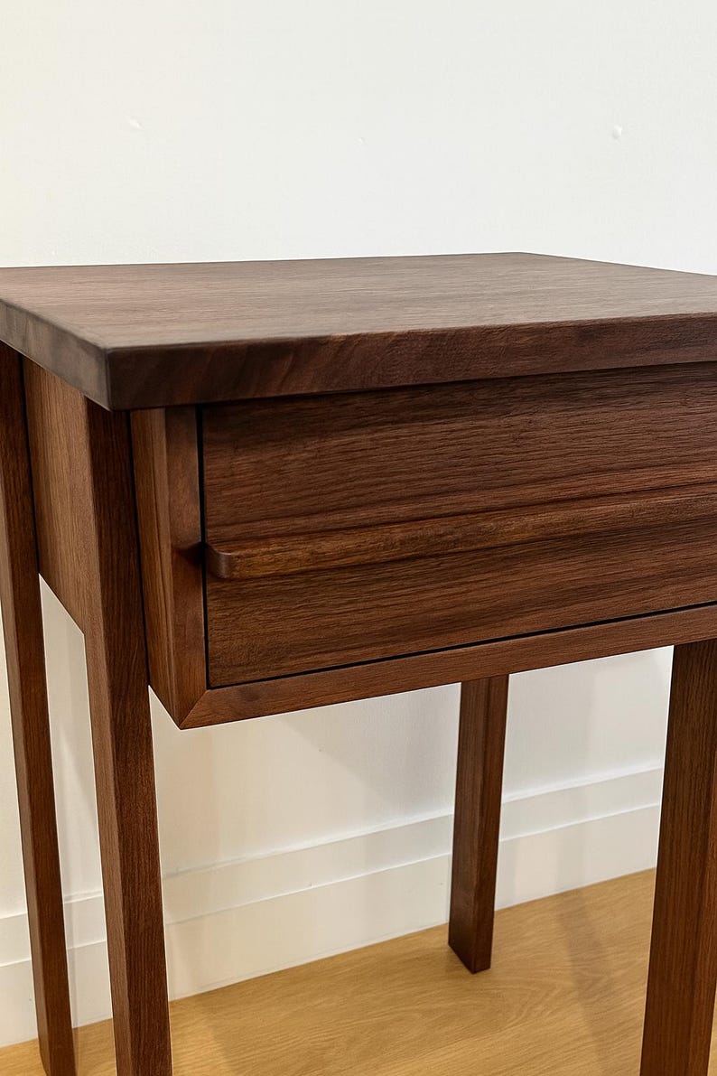 Pu&ograve; includere: Un tavolino in legno marrone scuro con un singolo cassetto. Il tavolo ha un piano rettangolare e quattro gambe sottili. Il cassetto presenta una maniglia semplice e curva. La venatura del legno &egrave; visibile.