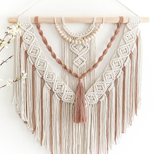 May include: A macrame wall hanging featuring a natural wood dowel and a complex design. The piece incorporates off-white and brown cords, with a central tassel and wooden bead accents. The geometric patterns and textures create a bohemian aesthetic.