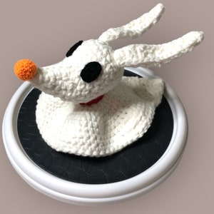 May include: A white crocheted ghost dog figure with long ears, black oval eyes, and a small orange nose. The figure features a red collar around its neck and a flowing, skirt-like base, crafted with visible yarn stitches in a textured, handmade style.