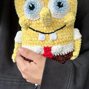 May include: A handmade, yellow crochet SpongeBob SquarePants plush toy. The toy features large blue and white eyes, a prominent nose, and a smiling mouth. It has a red tie, white shirt, and brown trousers. The toy is held in a person's hand.