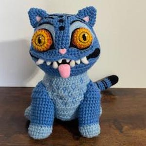 May include: A handmade blue crochet cat doll with large orange eyes, a pink tongue, and white teeth. The cat has a striped tail and is sitting upright. The doll is made of yarn and has a textured appearance.