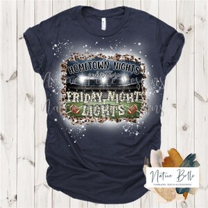 May include: A navy blue t-shirt with a white splatter design. The design features a football field with lights, two footballs, and the text "HOMETOWN NIGHTS under those FRIDAY NIGHT LIGHTS".