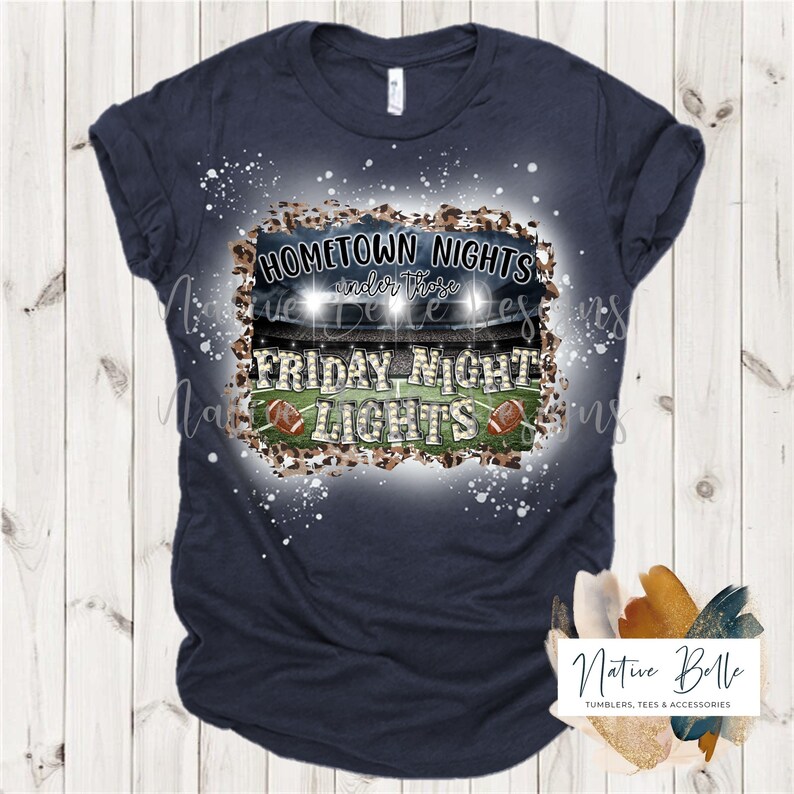 May include: A navy blue t-shirt with a white splatter design. The design features a football field with lights, two footballs, and the text "HOMETOWN NIGHTS under those FRIDAY NIGHT LIGHTS".