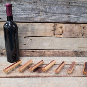 May include: A dark wine bottle with a burgundy top next to six wooden bottle stoppers. The stoppers are cylindrical with a light wood grain and a darker wood accent on the end. The items are on a rustic wooden surface.