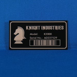 Peut inclure: Une étiquette rectangulaire noire avec du texte blanc et un logo de chevalier blanc. L'étiquette indique "KNIGHT INDUSTRIES" avec "Modèle: K2000" et "N° de série: AD227529" et un code-barres. Le fond est bleu.