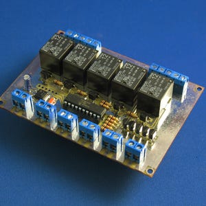 Op de afbeelding: Een printplaat met meerdere zwarte relais, blauwe klemmen en diverse elektronische componenten. De rechthoekige plaat heeft een metalen afwerking en is geplaatst tegen een blauwe achtergrond. De componenten zijn geordend.
