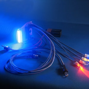 May include: A circuit board with wires and LEDs. The LEDs are lit in blue, white, and red. The circuit board is connected to a battery pack.