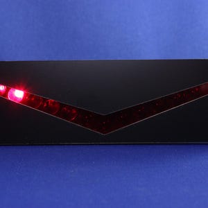 May include: A black rectangular device with a V-shaped red light design. The device has several red lights illuminated within the V-shape. A red cable extends from the right side of the device.