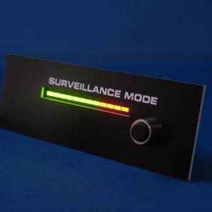 Peut inclure: Panneau rectangulaire noir avec le texte "SURVEILLANCE MODE" et un indicateur de barre horizontale. La barre passe du vert au jaune au rouge. Un petit bouton rond se trouve sur le côté droit du panneau.