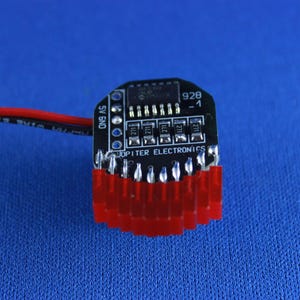 May include: A small black circuit board with red connectors at the bottom and a red and black wire extending to the left. The board has several small components and the text "JUPITER ELECTRONICS". The background is a solid blue.