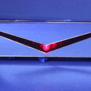 May include: A rectangular, mirrored object with a V-shaped red light. The object is mounted on blue supports and has a red wire extending from it. The background is a solid blue.