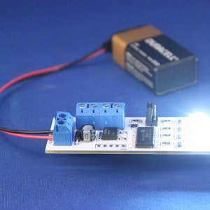 May include: A white circuit board with a blue connector, a black wire, and a red wire connected to a battery. A white LED light is lit on the board.