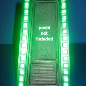 以下が含まれることがあります： 側面が明るい緑色のLEDライトが付いた緑色の電子デバイス。「pedal not included」というテキストがデバイスの前面に白で表示されています。デバイスは青色の背景にあります。