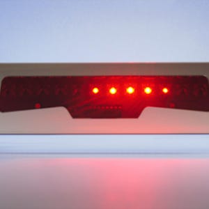 May include: A rectangular, red LED light bar with five illuminated bulbs. The light bar is set within a white frame, and the lights are bright against a light background. The product appears to be an automotive part.