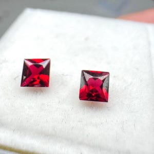 May include: Two square-cut, faceted, deep red gemstones. The gems are set against a white background. The facets create a brilliant sparkle, showcasing the rich color and clarity of the stones. These gems are likely for jewelry.