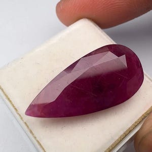 May include: A pear-shaped, faceted gemstone with a deep magenta-red color. The stone features a smooth, polished surface with multiple geometric facets that reflect light, showcasing its translucent quality and rich, saturated red hue.