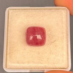 May include: A cushion-cut, translucent pink gemstone with a smooth, polished surface. The stone is centered on a square, off-white display pad with a thin gold-colored border, housed within a white plastic container.
