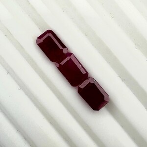 Peut inclure: Trois pierres précieuses rouge foncé, taillées en émeraude et disposées en ligne diagonale. Chaque pierre présente une surface à facettes avec des bords nets et une forme rectangulaire, affichant une teinte cramoisie sombre et uniforme.