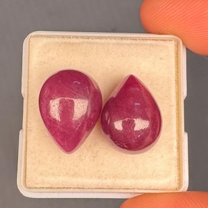 May include: Two pear-shaped, polished, deep pinkish-red gemstones resting on a square, cream-colored foam insert within a white plastic display box. The stones have a smooth, rounded surface with visible light reflections on their tops.