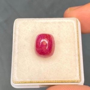 Könnte beinhalten: Ein leuchtender, tief pink-roter Edelstein mit einem glatten, abgerundeten Cabochon-Schliff. Der Stein hat eine polierte, reflektierende Oberfläche und ist mittig in einer quadratischen, weiß ausgekleideten Schaumstoff-Präsentationsbox platziert.