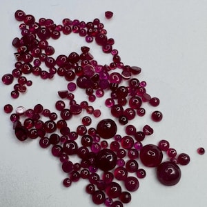 Può includere: Una collezione di gemme di rubino sciolte e lucide di varie dimensioni. Le gemme variano dal rosso intenso a tonalità più chiare di rosa, con una superficie lucida e riflettente. Le gemme sono sparse su uno sfondo bianco.