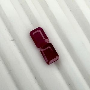 Könnte beinhalten: Zwei tiefrote, rechteckig geschliffene Edelsteine mit facettierten Oberflächen. Diese losen Steine weisen einen klassischen Smaragdschliff mit scharfen Ecken und einer polierten Oberfläche auf und zeigen einen satten, kräftigen Karmesinton.
