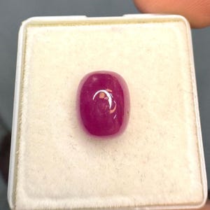 Könnte beinhalten: Ein leuchtender, tief pink-roter Edelstein mit einem glatten, abgerundeten Cabochon-Schliff. Der Stein hat eine polierte, reflektierende Oberfläche mit einem sichtbaren Lichtreflex und liegt mittig in einer quadratischen, weißen Box.