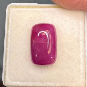 May include: A rectangular, cabochon-cut gemstone with a deep magenta-pink color and a smooth, polished surface. The stone features subtle internal inclusions and a reflective white light highlight on its top center, set in a white display box.
