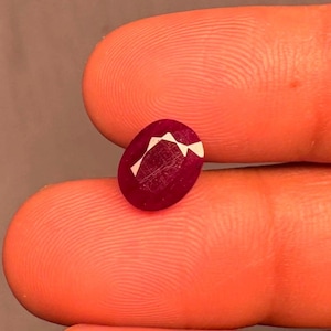 Könnte beinhalten: Ein tiefroter, oval geformter Edelstein mit einer glatten, polierten Oberfläche und einer markanten weißen, kantigen Reflexion im oberen Bereich. Der Stein zeigt einen satten, kräftigen Karmesinton und wird zwischen zwei Fingern gehalten.