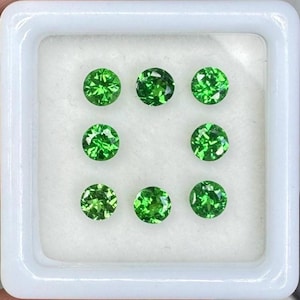 May include: Nine round, faceted, bright green gemstones are arranged in a square within a white square container. The gems have a brilliant, sparkling appearance, suggesting high clarity and quality. The gemstones are likely for jewelry making.