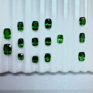 May include: A collection of vibrant green gemstones, each with a unique cut and shape, arranged on a white surface. The gems vary in size and display a rich, emerald hue, reflecting light. The arrangement highlights the variety of the gemstones.