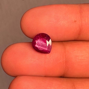 Peut inclure: Une pierre précieuse à facettes en forme de poire d'une couleur rouge magenta profonde et vibrante. La pierre présente une surface polie avec des inclusions internes visibles et des facettes géométriques nettes qui reflètent la lumière.