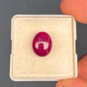 以下が含まれることがあります： 深いマゼンタレッドの色合いと目に見える暗いインクルージョンを持つ、磨かれた楕円形のルビーの宝石。保護と展示のために、柔らかい質感のクリーム色のフォームインサートを備えた正方形の白いディスプレイボックスの中央にしっかりと配置されています。