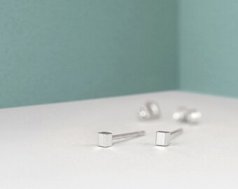 May include: A pair of silver square stud earrings on a white surface.
