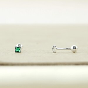May include: Two silver earrings. One features a square, emerald-green gemstone. The other is a barbell style earring with a small ball on one end and a flat, rectangular design on the other. The earrings are displayed on a light beige surface.