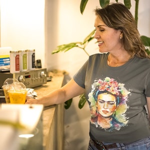 May include: A woman wearing a gray t-shirt with a colorful watercolor portrait of Frida Kahlo. The portrait features a floral headband and a colorful background.