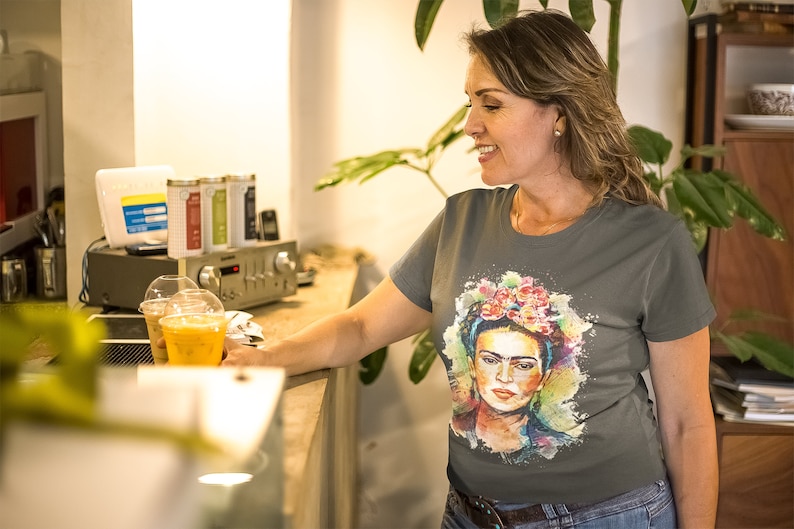 May include: A woman wearing a gray t-shirt with a colorful watercolor portrait of Frida Kahlo. The portrait features a floral headband and a colorful background.
