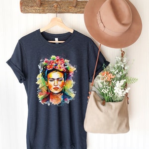 May include: A dark blue t-shirt with a colorful watercolor portrait of Frida Kahlo. The portrait features her signature flower crown and a colorful background.
